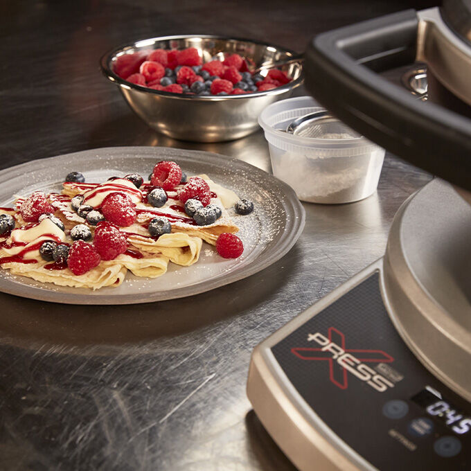  Work surface with a crepe maker, a plate of folded crepes with berries, and a bowl of berries.